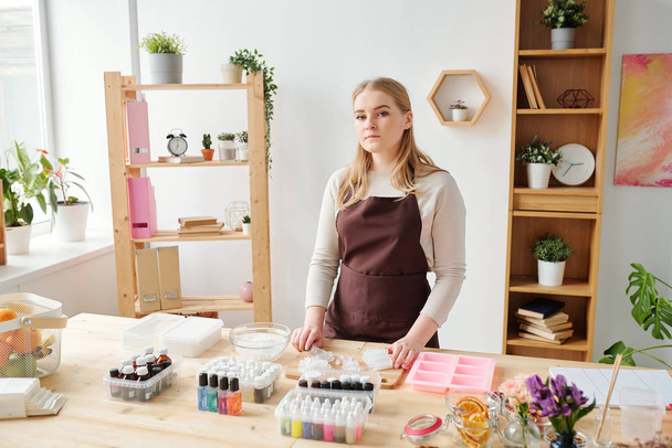 junge blonde Handwerkerin in brauner Schürze steht im Atelier am Tisch, während sie handgemachte Seife herstellt - Foto, Bild
