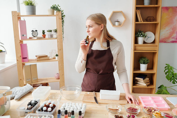 Young blonde female in brown apron smelling aromatic essence in small bottle while choosing scent for soap - Photo, Image