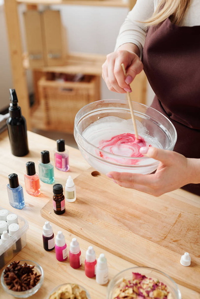 Hands of craftswoman mixing liquid soap mass with crimson color in glassware with wooden stick over table - Photo, Image