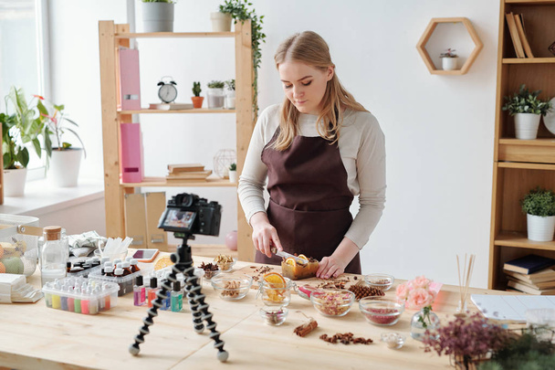 Young craftswoman shooting home master class on camera while standing by table and cutting large handmade soap bar - Photo, Image
