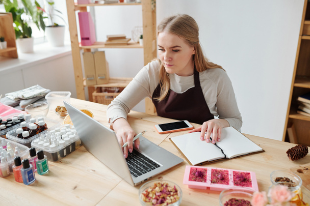 Pretty młoda kreatywna kobieta w fartuchu siedzi w miejscu pracy przed laptopem robiąc notatki o jej hobby - Zdjęcie, obraz