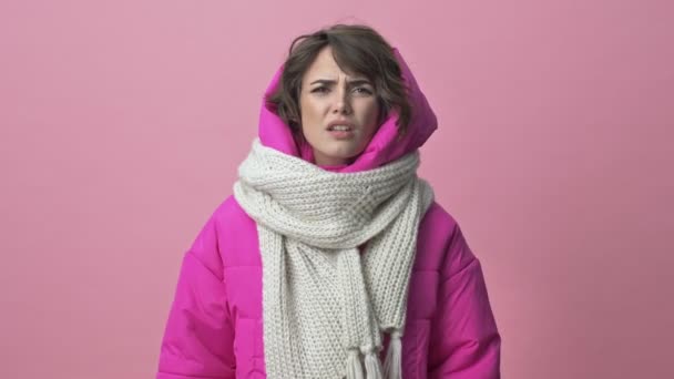 Puzzled young woman wearing a winter jacket with a scarf looking confused while shrugging her hands isolated over a pink background - Filmati, video