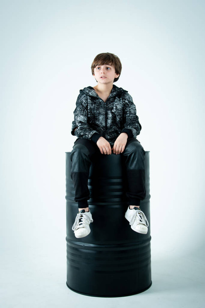 boy in black zip hoodie sitting on black barrel on gray background - Φωτογραφία, εικόνα