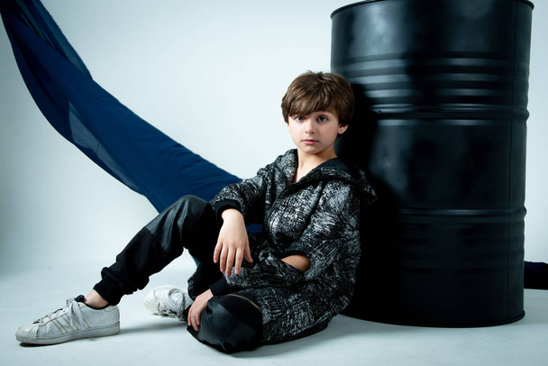 boy in black zip hoodie sitting near black barrel on gray background - Zdjęcie, obraz