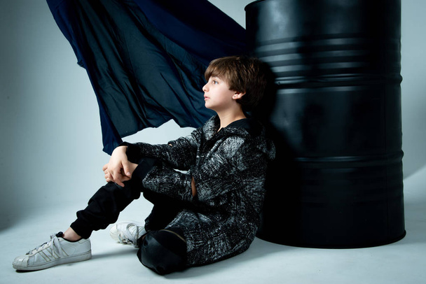 brunet kid boy sitting with black barrel on gray background - Фото, изображение