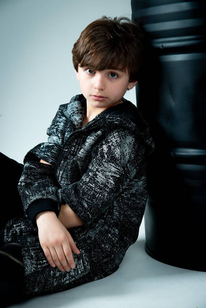 brunet kid boy sitting with black barrel on gray background - Φωτογραφία, εικόνα