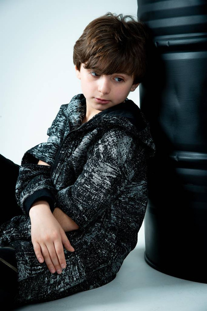 brunet kid boy sitting with black barrel on gray background - Photo, Image