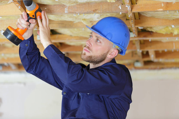 young builder drilling wooden ceiling joist - Fotó, kép