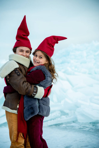 smiling children in red gnome caps hugging on ice on winter background - Photo, Image