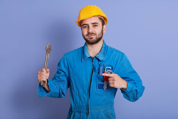 Close up portrait of bearded engineer with wrenches looking at camera and pointing athis tool with index finger, repairman posing against blue background, mechanic dresses uniform and yellow hard hat. - Φωτογραφία, εικόνα