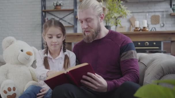 Cute Caucasian girl showing something in book and shaking head no. Young father reading for daughter at home. Good relationship, family, care, hobby, lifestyle. - Filmati, video
