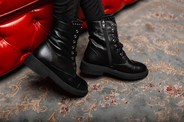 Close up of female legs in trendy jeans in seasonal stylish leather black lace-up boots in the room. Modern girl in fashionable shoes sits on a red soft chair in the store. Women's winter footwear. - Fotó, kép