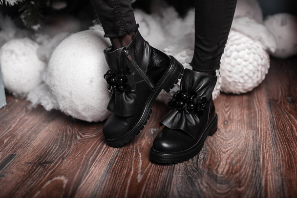 Стильні красиві чорні шкіряні зимові черевики на жіночих ногах біля білих вінтажних іграшок. Новорічний розпродаж. Модне зимове жіноче взуття. Крупним планом
. - Фото, зображення