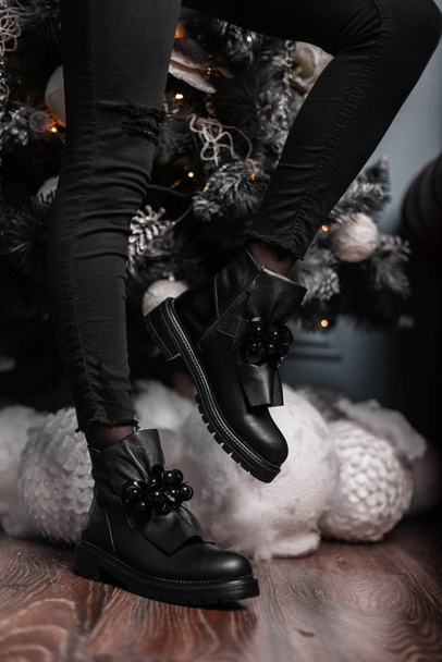 Mujer joven de moda en jeans vintage en zapatos negros de cuero de moda posa cerca de un árbol de Navidad en la habitación. Primer plano de las piernas femeninas en botas con estilo. Calzado juvenil de moda de invierno
. - Foto, Imagen