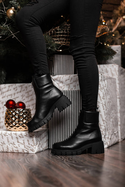 Kerst verkoop schoenen. Winter collectie van warme leren damesschoenen. Close-up van vrouwelijke benen in zwarte trendy laarzen in de buurt van feestelijke geschenken en spiegelballen. - Foto, afbeelding