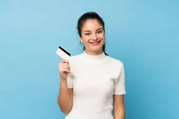Young brunette girl over isolated blue background holding a credit card - Zdjęcie, obraz