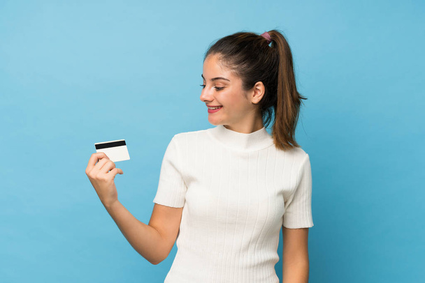 Young brunette girl over isolated blue background holding a credit card - Фото, изображение