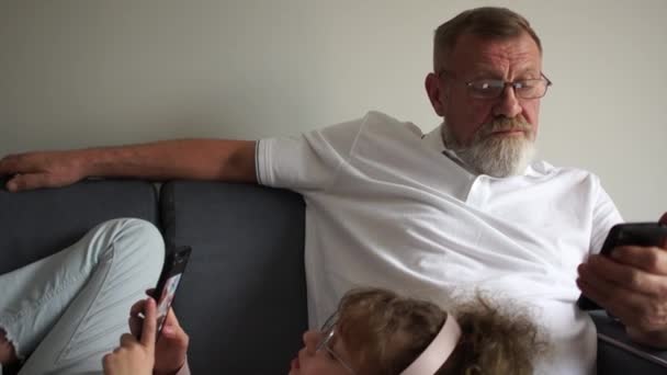 Granddaughter teenager resting on the couch, lying on his grandfathers lap. Grandfather and granddaughter look at the screens of their smartphones, family and smartphones - Metraje, vídeo