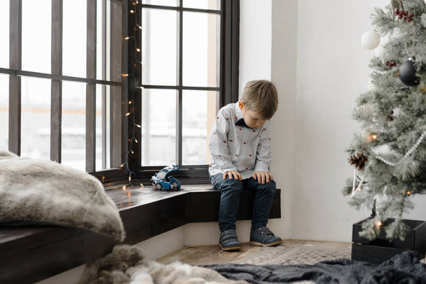 Offended little boy sitting next to a beautiful dressed-up Christmas tree - Фото, изображение