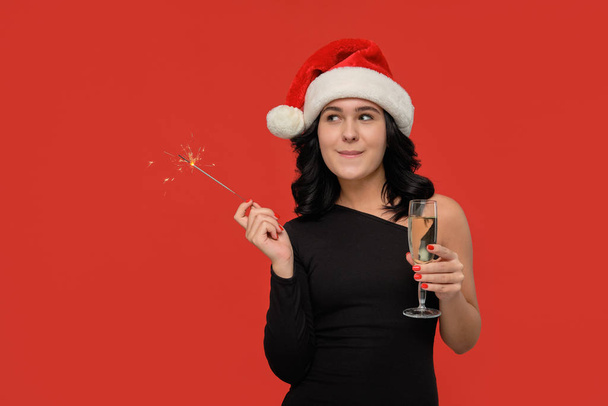 Beautiful brunette girl in a black dress and Santa hat holding glass of champagne and bengal lights. - Photo, Image