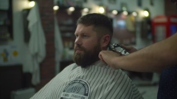 Barber making haircut for beard man in hairdresser cape by electric shaving machine and comb in barbershop in slow motion.  - Záběry, video