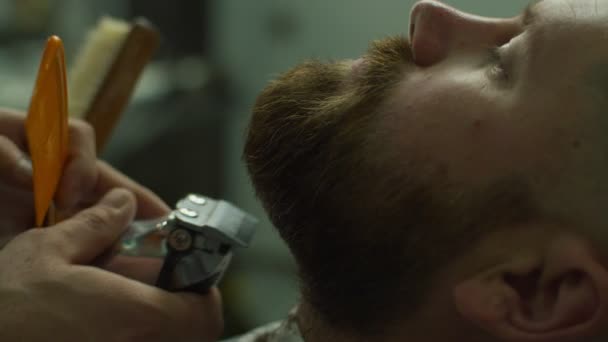 Bearded man and barbers hands shaving beard by electric shaving machine and comb in slow motion. Male with closed eyes in barbershop.  - Záběry, video