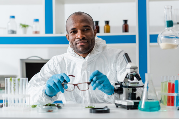 smiling african american biologist holding glasses and looking at camera  - Foto, afbeelding