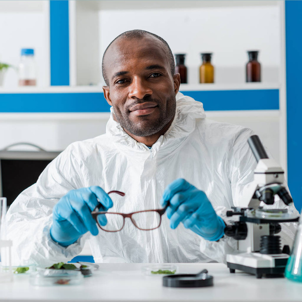χαμογελαστός αφροαμερικανός βιολόγος κρατώντας γυαλιά και κοιτάζοντας την κάμερα  - Φωτογραφία, εικόνα