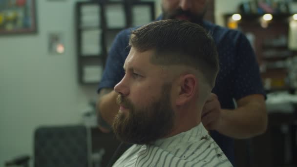 Side view of barber making haircut for man in hairdresser cape in barbershop in slow motion.  - Video, Çekim