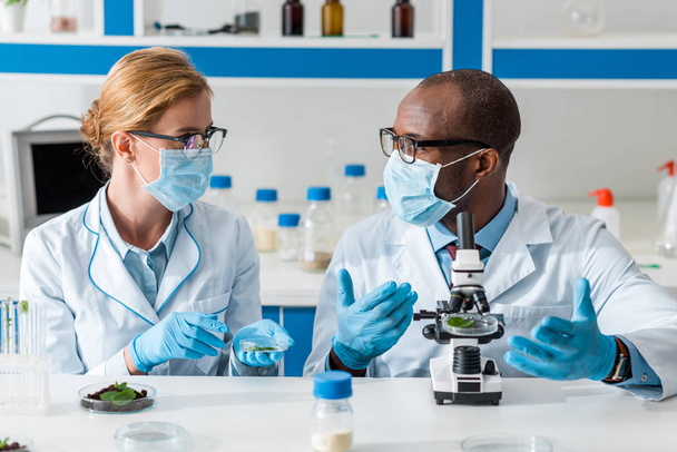 biólogos multiculturales con batas blancas y máscaras médicas hablando en laboratorio
  - Foto, Imagen