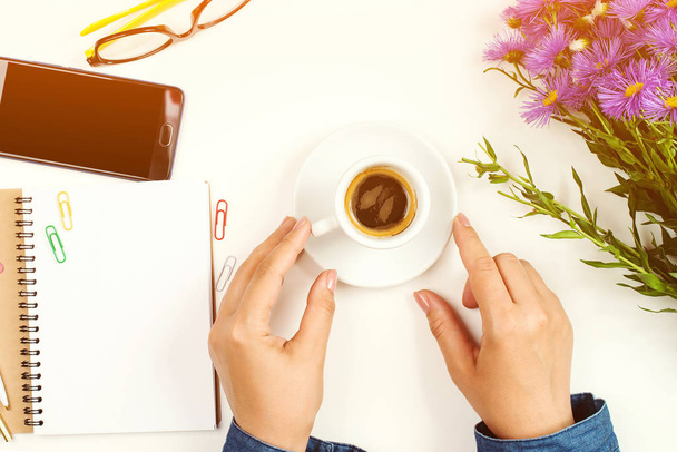 Kadın eli beyaz ofis masasında bir fincan kahve tutuyor. Masada temiz defter, gözlük ve bahar çiçeği var. - Fotoğraf, Görsel