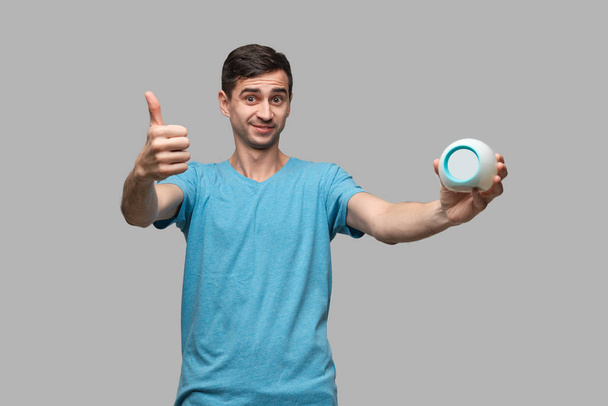 Young man showing thumb up holding alarm isolated over grey background. Concept of being in time. - Photo, Image