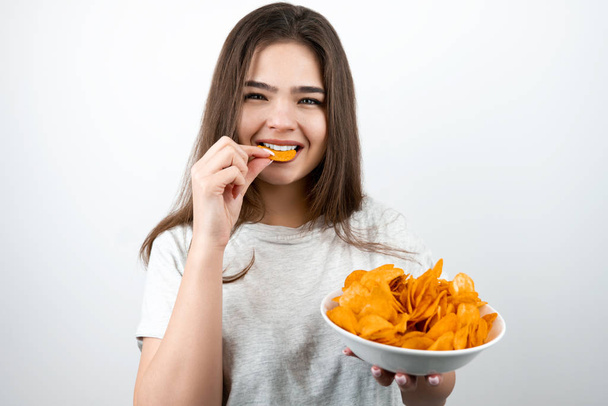 young attractive woman putting one piece of chips in her mouth standing with plate with spicy potatoe chips on isolated white background dietology and nutrition - Foto, imagen