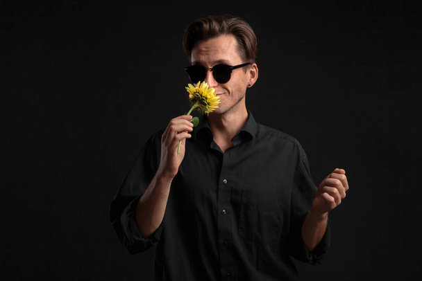 Przystojny mężczyzna w czarnej koszuli i okularach przeciwsłonecznych trzymający żółty kwiat, by dać go ukochanemu.. - Zdjęcie, obraz