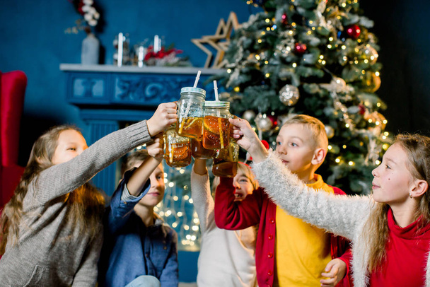 Group of children on the background of a Christmas tree , drinking lemonade and knocking glasses, focus on glasses - Photo, image