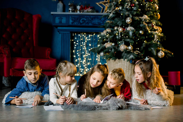 Small charming and beautiful children write a letter to Santa in the background of a Christmas tree on the eve of the holidays - 写真・画像