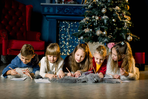um grupo de crianças pequenas jazem em um cobertor de lã quente no fundo de uma árvore de Natal e escrevem uma carta para o Papai Noel
 - Foto, Imagem