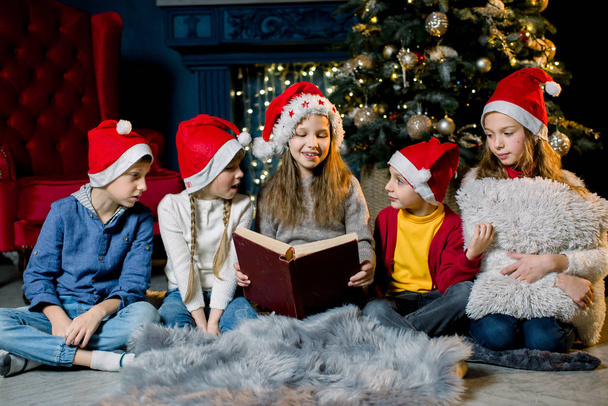 On the eve of Christmas, beautiful little children look at old book with Christmas stories in a warm cozy room in red New Year hats - Foto, Imagem