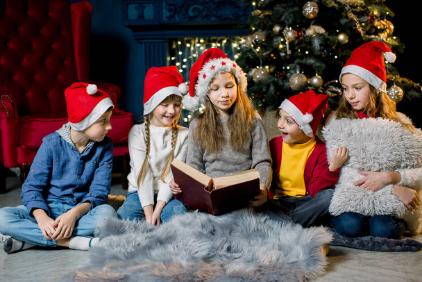 Beautiful little children read a book with Christmas stories on the background of a Christmas tree in red New Year hats - Zdjęcie, obraz