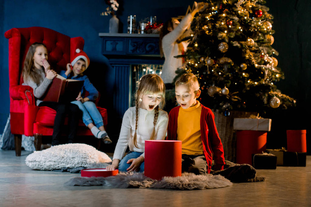 Beautiful little children look at a magical gift, their brothers and sisters read a book with Christmas stories and decorate a Christmas tree. - Фото, изображение