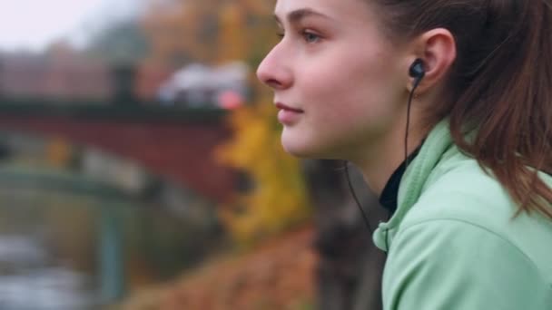 Tracking around shot of beautiful sporty girl in earphones dreamily resting after running in city park on riverside - Materiał filmowy, wideo