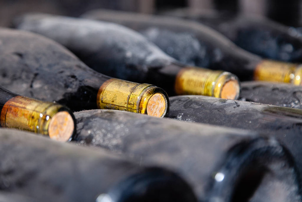 Στοίβες φιαλών κρασιού ξαπλωμένες σε ράφια σε παλιό κελάρι ή σπηλιά. Κονσόλα με παλιά σπάνια κρασιά, αποκλειστική συλλογή. Αρχαία μπουκάλια κρασιού που αναπαύονται, γερνούν, ξεσκονίζουν στο κελάρι - Φωτογραφία, εικόνα