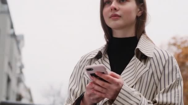 Стежити за зйомками красивої випадкової дівчини в траншеї пальто мрійливо, використовуючи мобільний телефон на міській вулиці
 - Кадри, відео