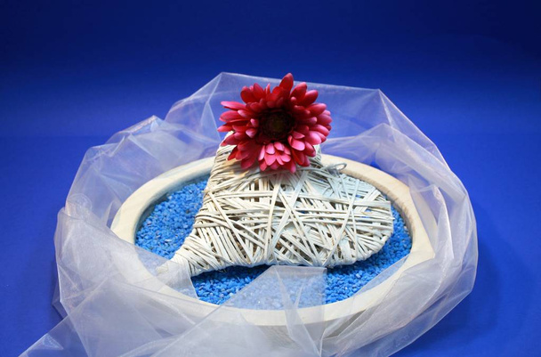 Coração de bast com crisântemo de algodão vermelho em pedras decorativas azuis em tigela de madeira
 - Foto, Imagem