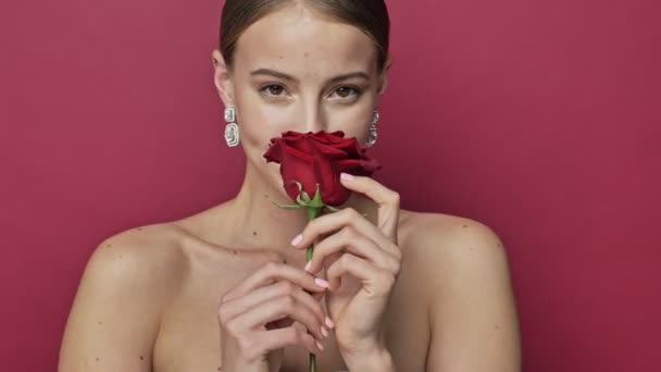 Beautiful young woman with red lipstick and earrings is poising with a red rose flower isolated over red background - Filmati, video