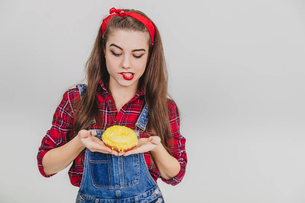 Lovely young female holding tasty big doughnut with yellow icing in her hands, twisting her lip enticingly. - Fotoğraf, Görsel