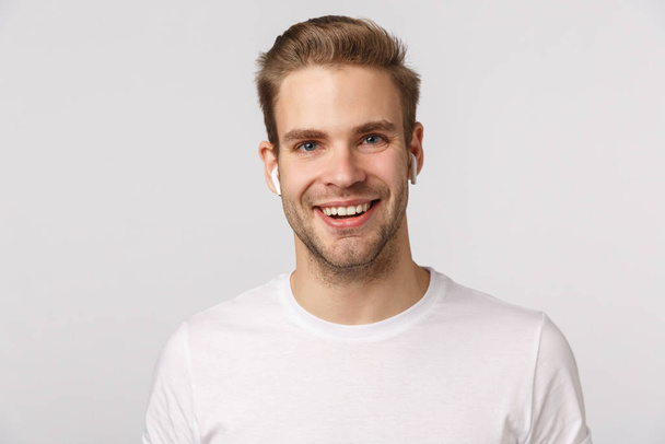 Handsome dreamy and excited blond caucasian guy with sexy bristle, wear wireless headphones and smiling, speaking, talking on phone using earphones, listen music earbuds, enjoy podcast - Valokuva, kuva