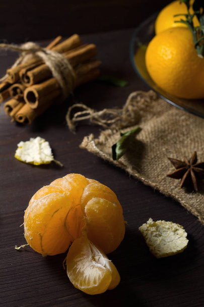 low key. peeled mandarin on the wooden table. fresh orange tangerines with green leaves on wooden background. ripe fruits on dark table with sticks of cinnamon and spices. - Photo, Image