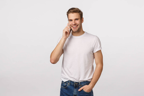 Business, technology and modern lifestyle concept. Handsome carefree smiling macho man with blond hair, bristle, casually talking on phone, hold smartphone near ear, grinning camera, white background - 写真・画像
