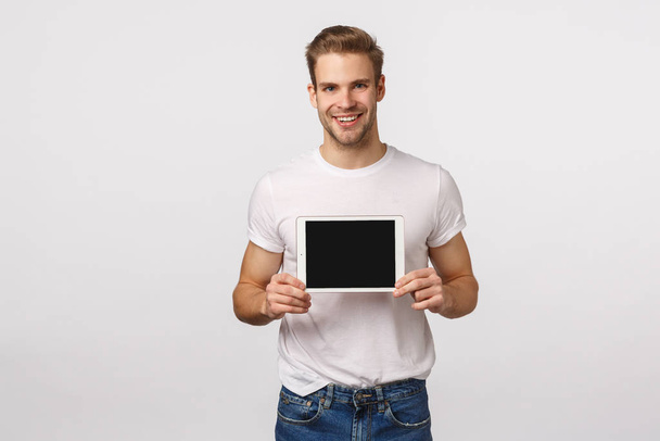 Must have, check this out. Attractive sassy caucasian blon bearded man in white t-shirt, holding digital tablet, showing device screen, introduce application, smiling, showing favorite shopping site - Fotografie, Obrázek